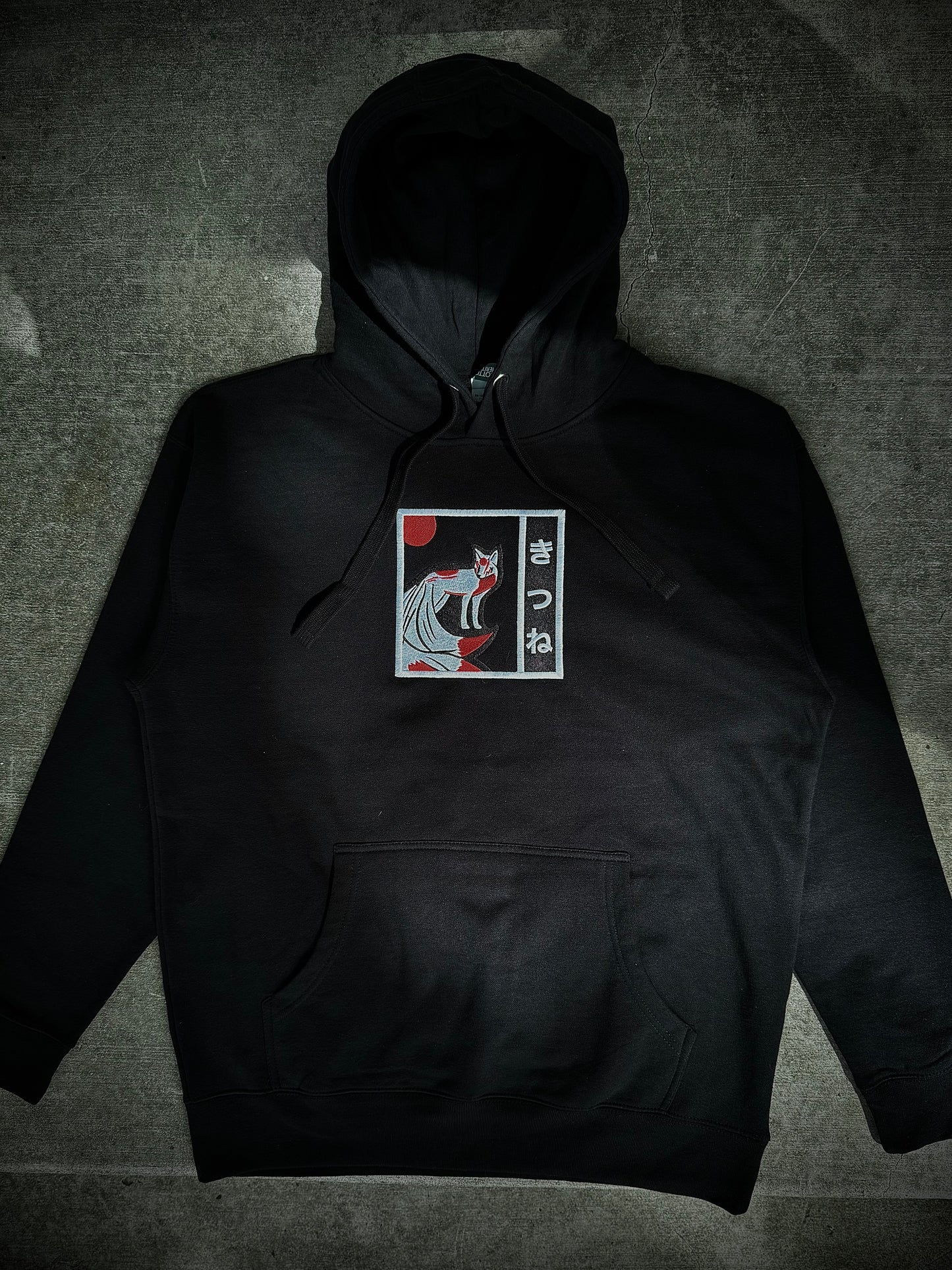 Flat lay of Kitsune embroidered hoodie on gray concrete background, showing black hoodie with minimalist white and red yokai embroidery on front.