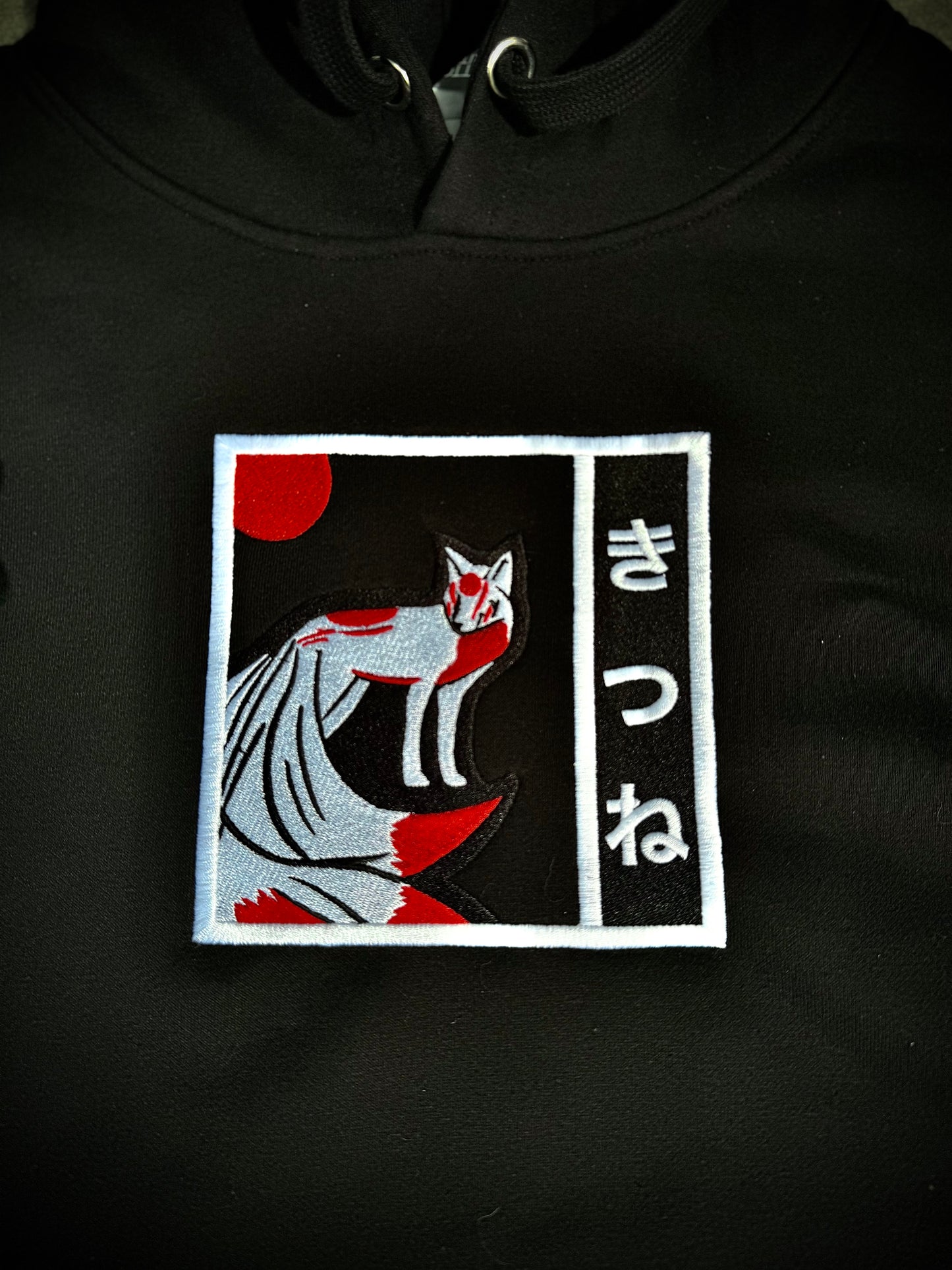 Close-up detail of Kitsune yokai embroidery on black hoodie, featuring balck, white, and red thread.