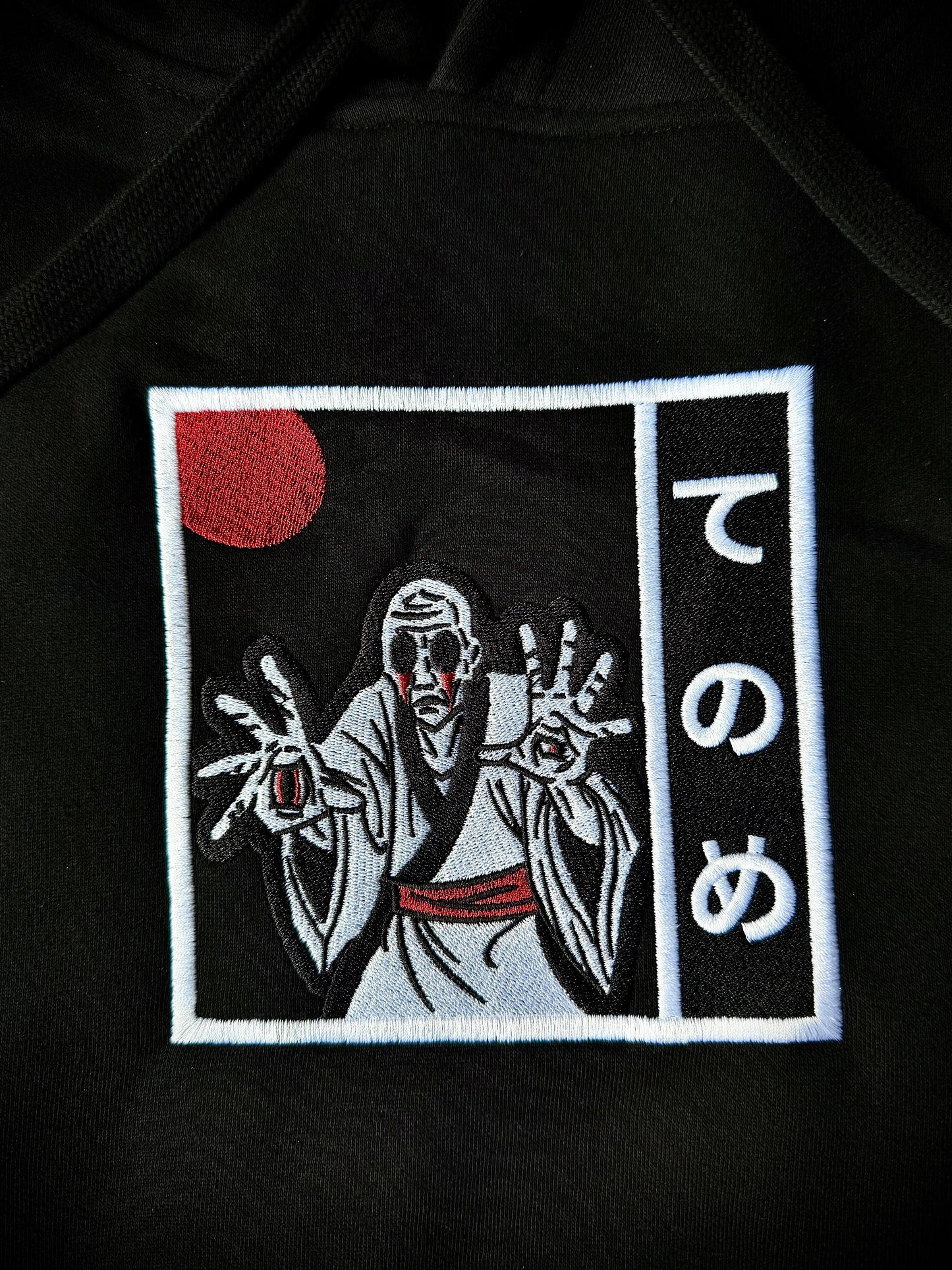 Close up of black streetwear hoodie showing black, white, and red embroidered Japanese yokai design of Tenome