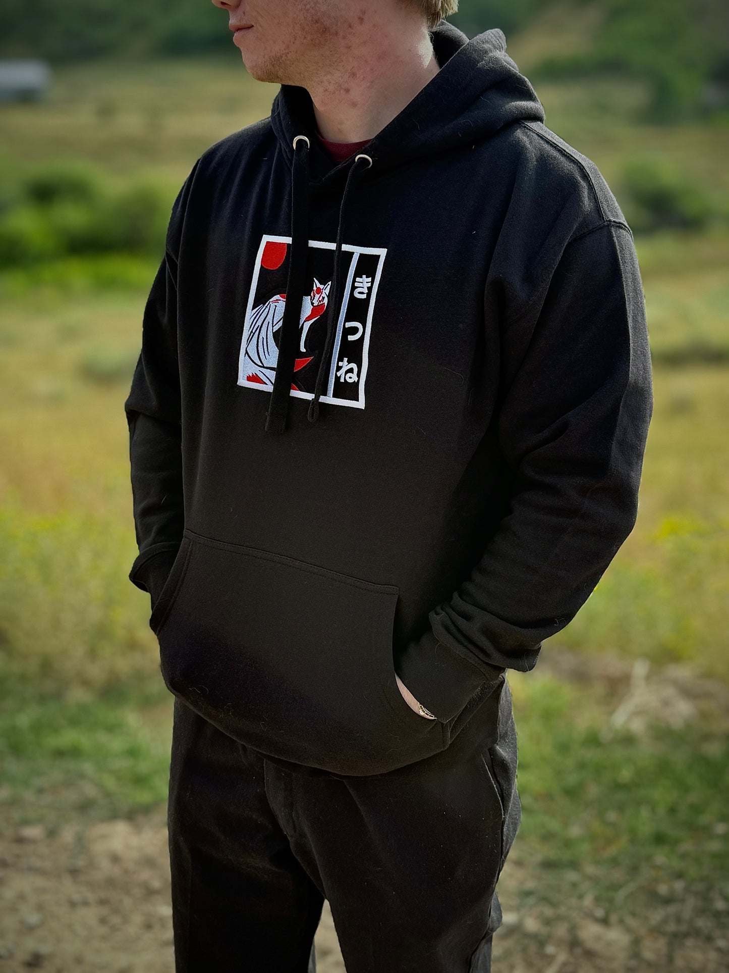 Person wearing black streetwear hoodie in nature, featuring embroidered Kitsune yokai design on the front.