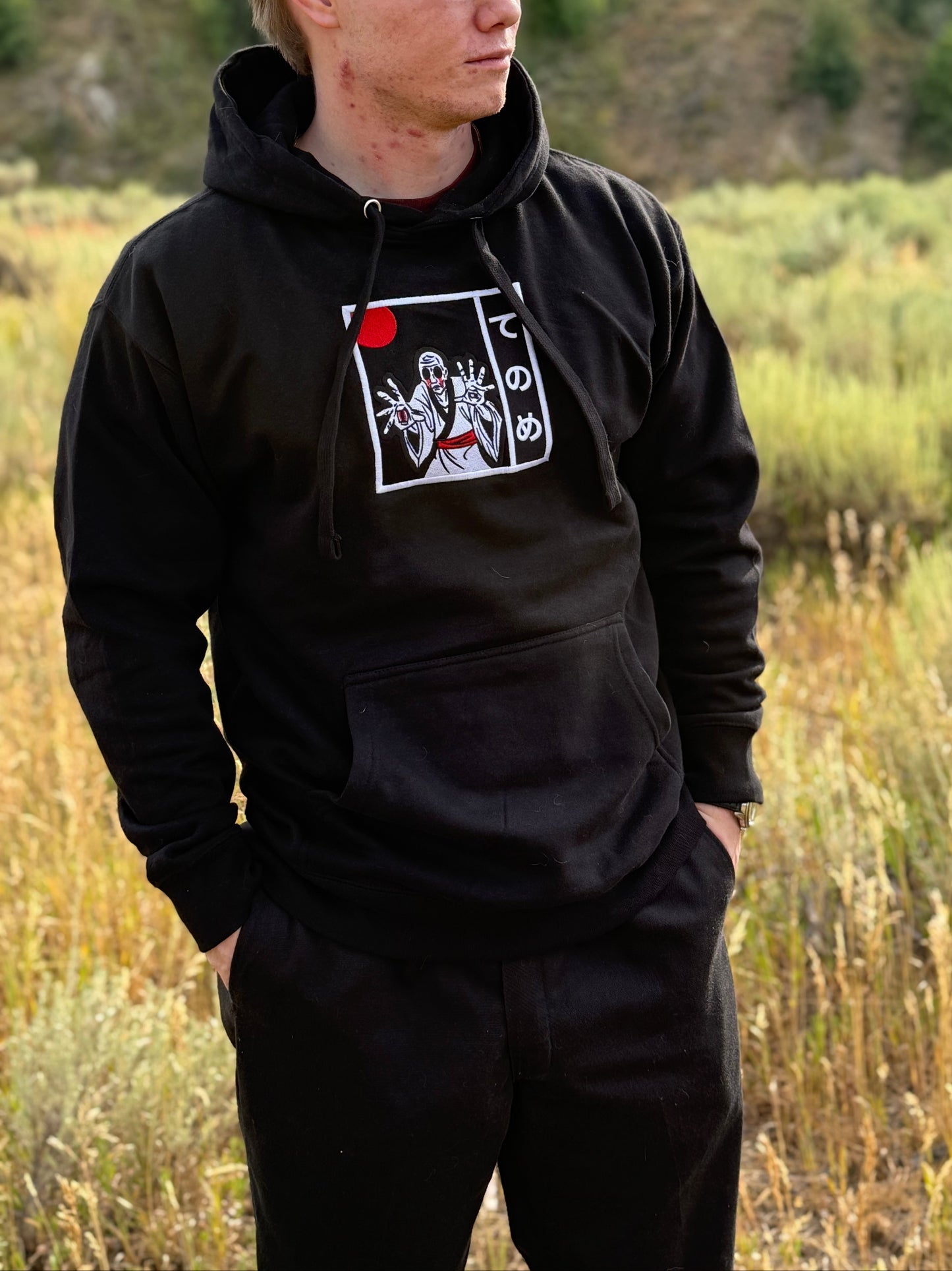 Person standing in nature, wearing black Japanese streetwear hoodie with embroidered Tenome yokai design on front.