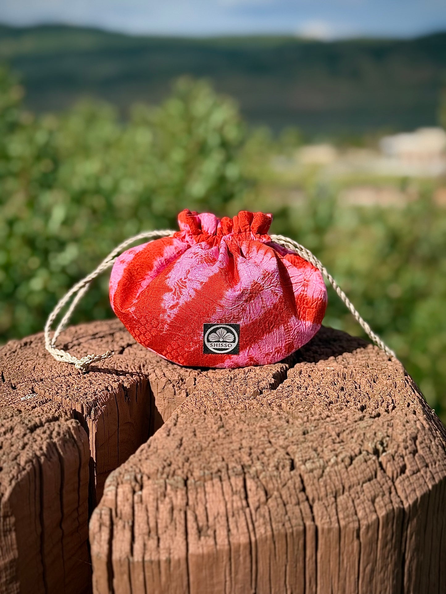 Red and pink kimono shibori silk drawstring bag featuring crepe and ripple textures with hemp drawstrings resting on top of a wooden post in nature