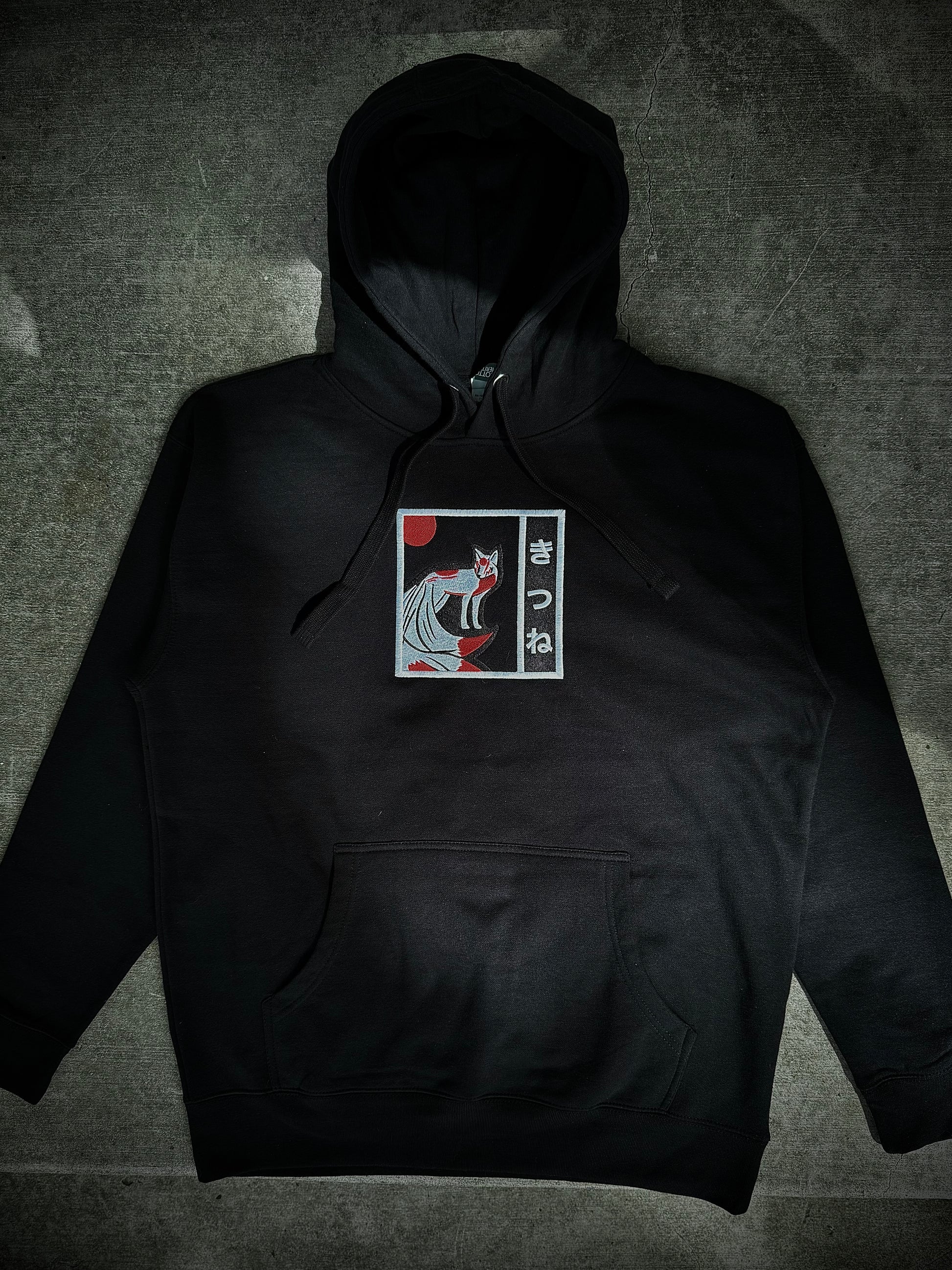 Flat lay of Kitsune embroidered hoodie on gray concrete background, showing black hoodie with minimalist white and red yokai embroidery on front.