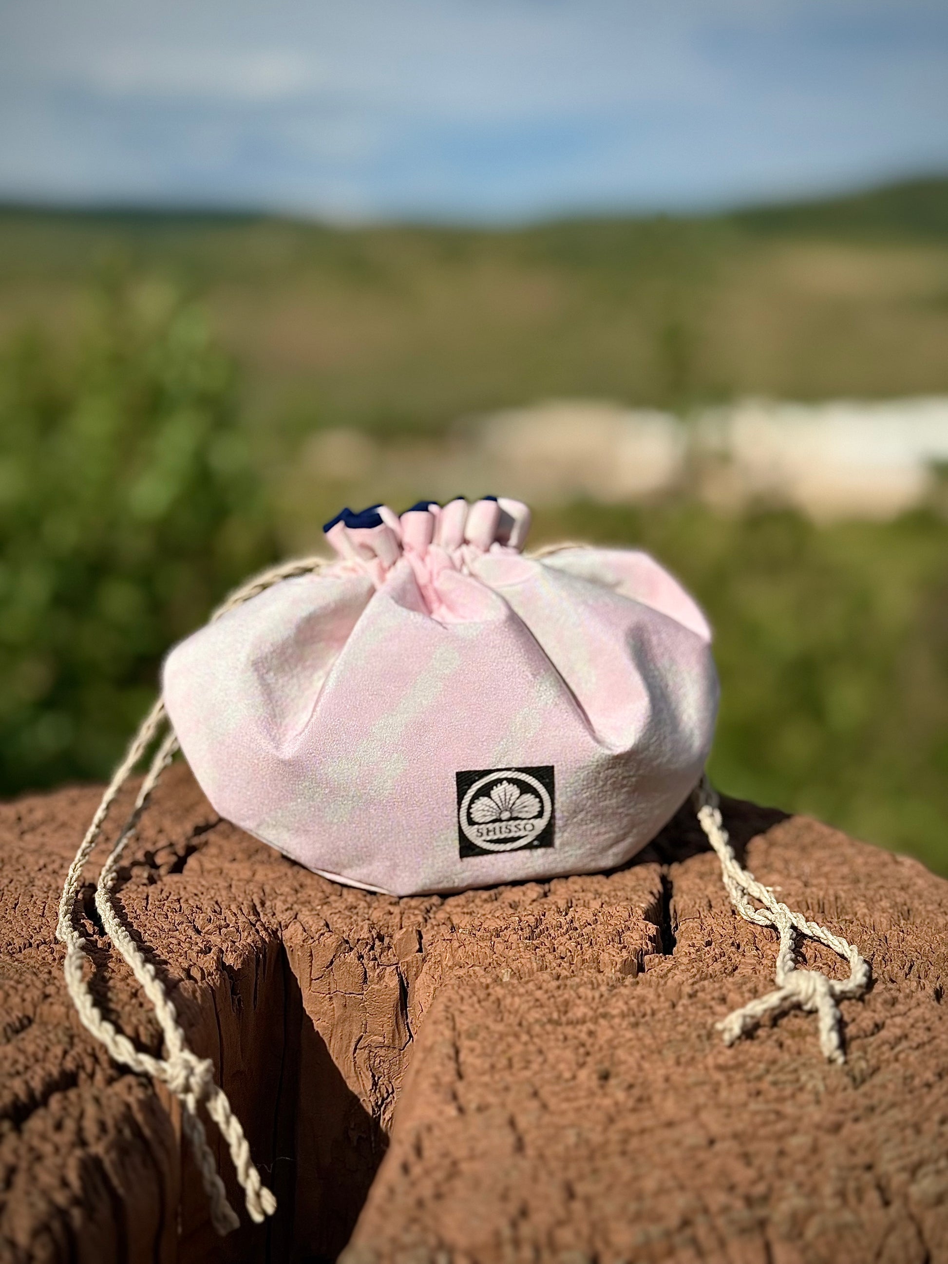 Light pink kimono silk drawstring pouch with subtle darker pink accents and hemp drawstrings cinched resting on a wooden post in nature