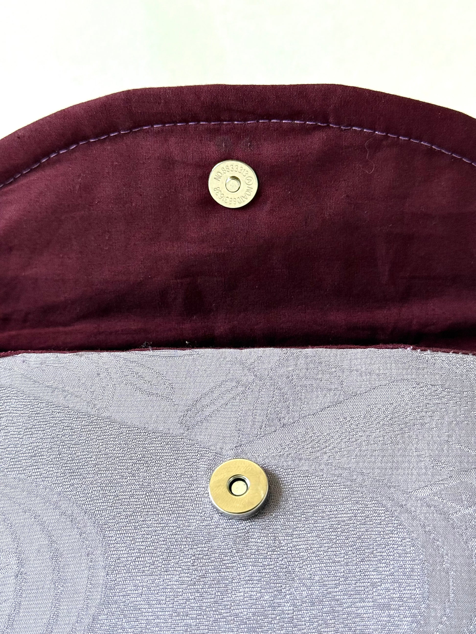 Close up of silver colored magnetic closure on wine colored cotton interior of top flat and main body of purple silk clutch 